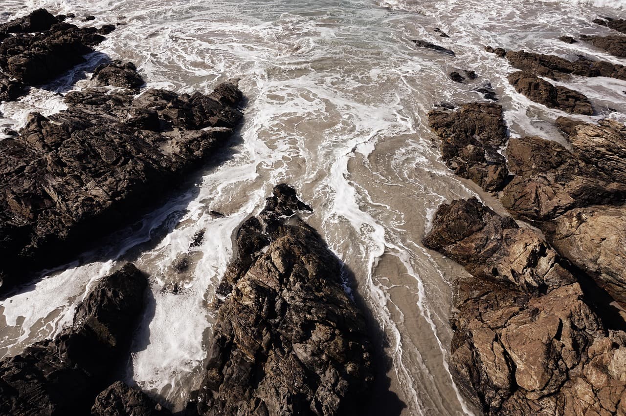 Olas rompiendo entre rocas sobre la arena bajo un cielo azul, en el chat de Carballo