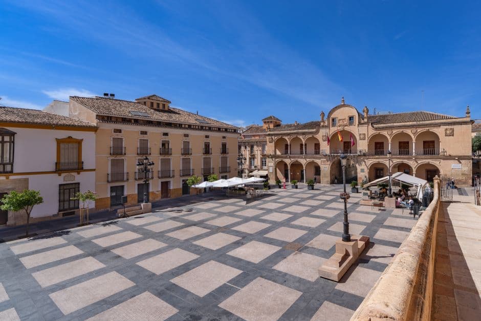Plaza soleada con arquitectura histórica y fuentes, en la sala de chat de Moguer