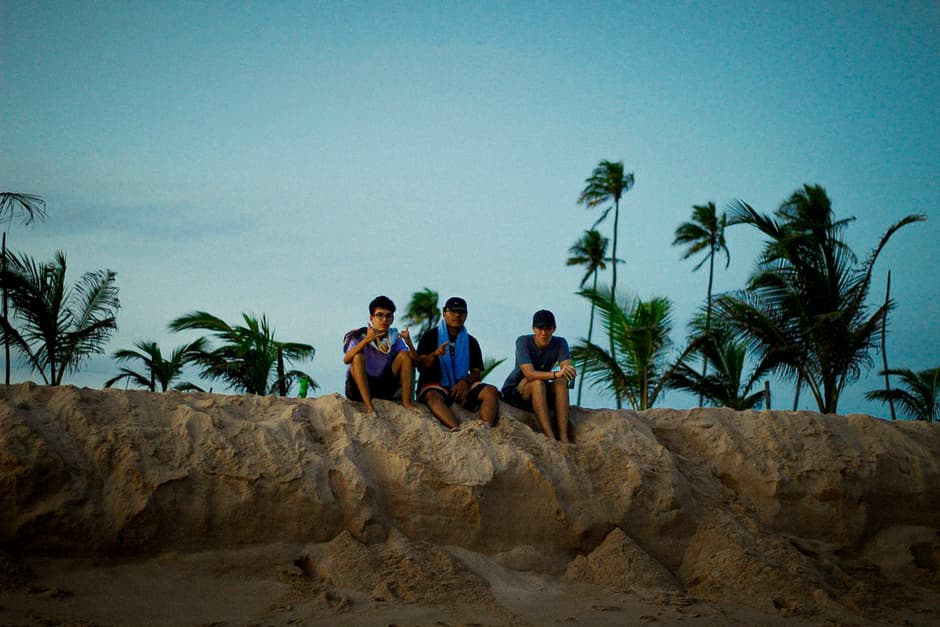 Hombres sentados en la arena bajo palmeras, mirando al horizonte, en el chat de Arapiraca