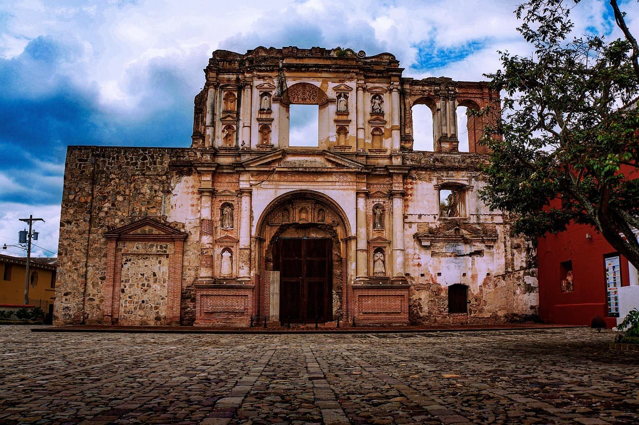 Iglesia colonial de piedra bajo cielo azul con vegetación, en la sala de chat de Cuyotenango