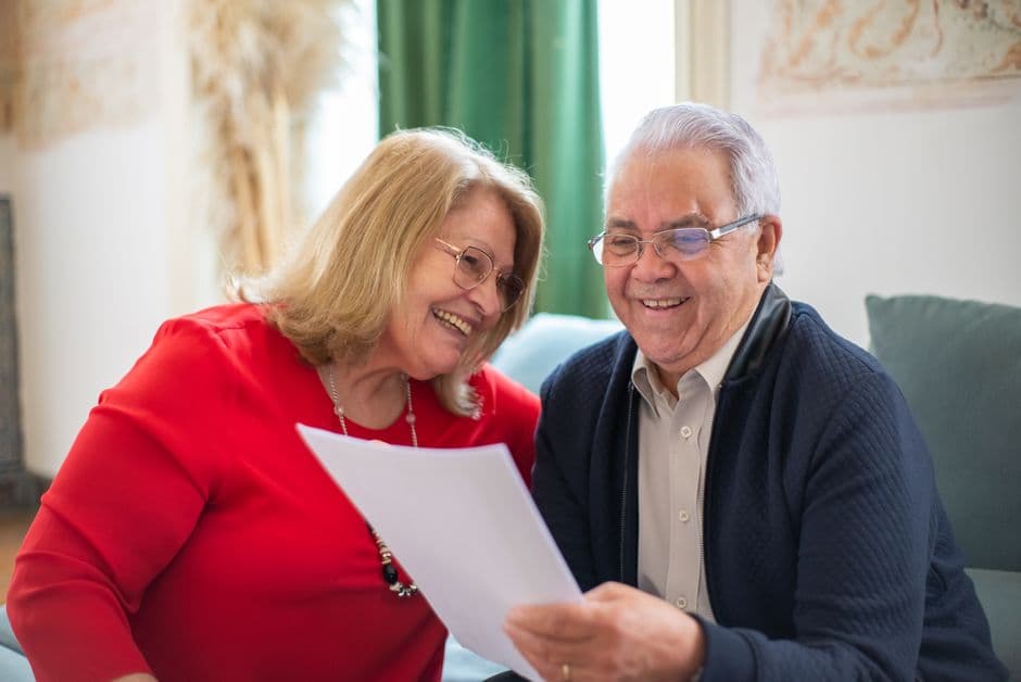 Pareja sonriente sentada en un sofá leyendo un papel, en la sala de chat de De 60 a 70